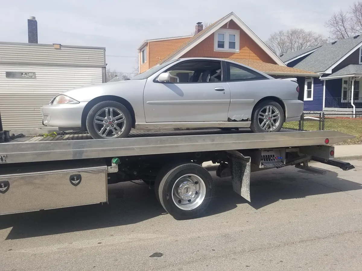 Rusted silver Chevy Cavalier coupe on a flatbed, a compact junk car at the low end of junkyard prices